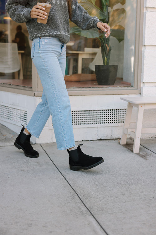 A person wearing light blue jeans, black boots, and a gray sweater walks on a sidewalk while holding a cup of coffee, passing by a window with a potted plant inside.