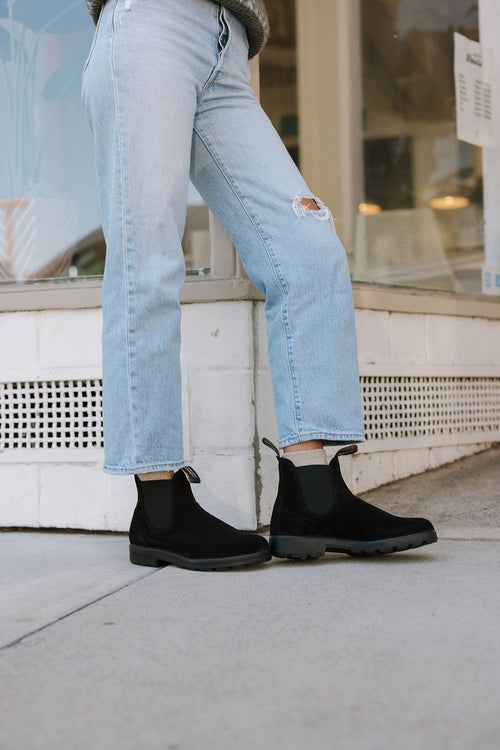 Person wearing light blue ripped jeans and black boots, standing on a sidewalk and leaning against a white wall outside a building with large windows.