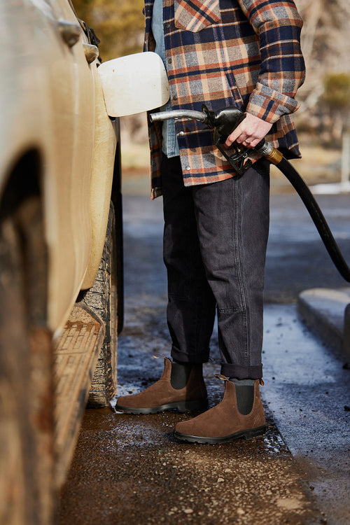 A person wearing a plaid jacket, dark pants, and brown boots is standing at a gas station, holding a fuel pump nozzle and refueling a dirty, light-colored vehicle. Only the lower half of the body is visible.