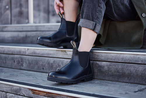 A person sits on wooden steps, wearing rolled-up dark jeans and shiny black Chelsea boots, preparing to stand up or adjust their boots.