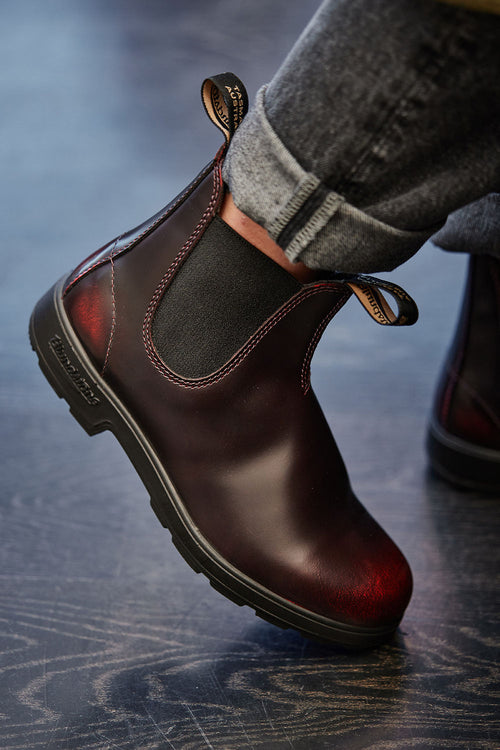 A person wearing rolled-up jeans and a dark brown Chelsea boot with black elastic sides, raising their foot slightly off a dark floor.