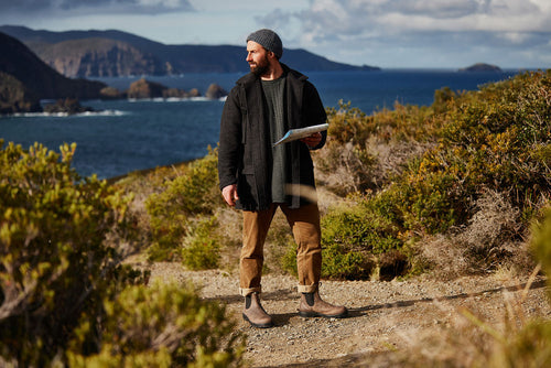 A bearded man wearing a gray beanie and dark coat stands on a dirt path by the coast, holding a map, surrounded by shrubs with the ocean and rocky islands in the background.