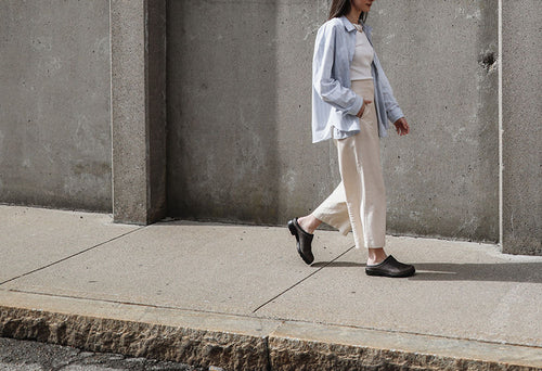 A person wearing a light blue shirt, white top, and cream pants walks along a concrete sidewalk next to a gray wall.