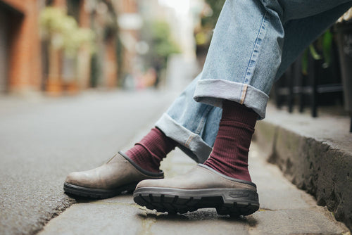 Person wearing cuffed blue jeans, burgundy socks, and brown slip-on clogs is sitting on a curb or ledge in an urban street setting, with their legs crossed at the ankles.